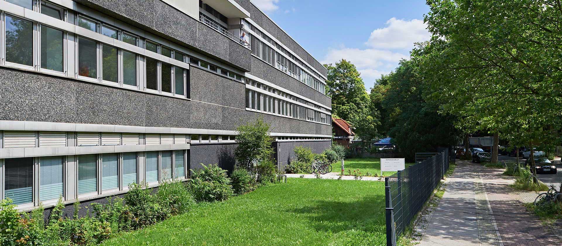 Modernes Laborgebäude des IHP mit grauer Fassade und großen Fenstern, umgeben von Bäumen und Rasen bei Sonnenschein.