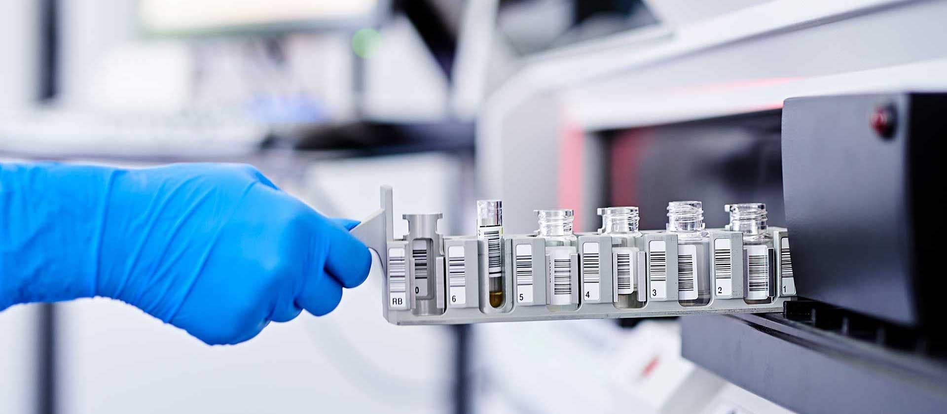Ein Laborant mit blauen Handschuhen platziert eine Reihe von Probenfläschchen mit Barcode-Etiketten in ein Analysegerät.