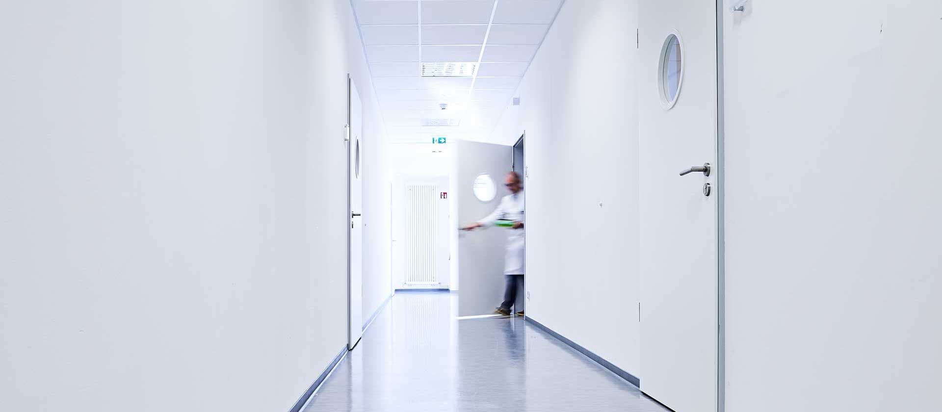 Ein heller, leerer Laborflur mit weißen Wänden und Türen. Im Hintergrund ist eine unscharfe Person in einem weißen Kittel zu sehen.