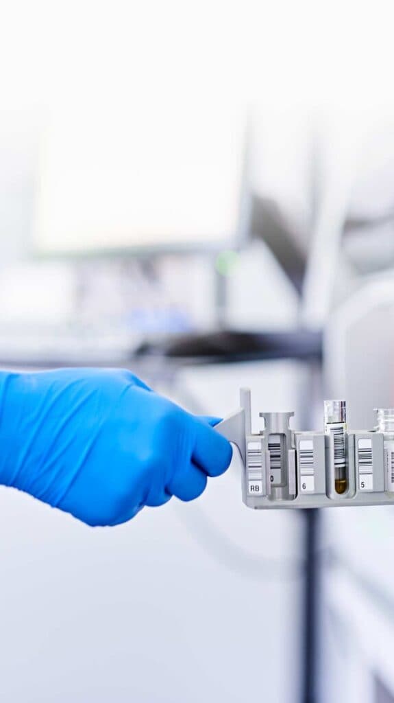 Ein Laborant mit blauen Handschuhen platziert eine Reihe von Probenfläschchen mit Barcode-Etiketten in ein Analysegerät.
