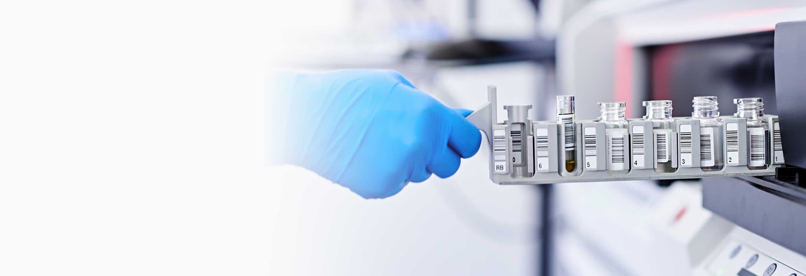 Ein Laborant mit blauen Handschuhen platziert eine Reihe von Probenfläschchen mit Barcode-Etiketten in ein Analysegerät.