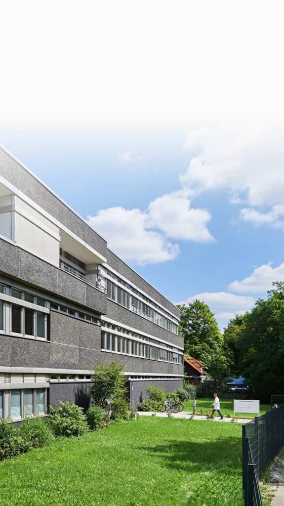 Modernes Laborgebäude des IHP mit grauer Fassade und großen Fenstern, umgeben von Bäumen und Rasen bei Sonnenschein.