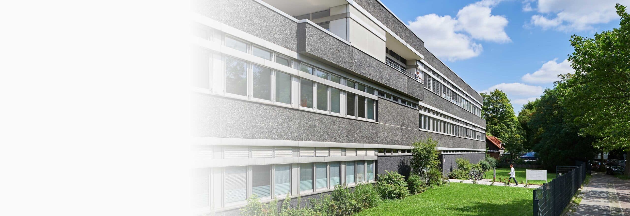 Modernes Laborgebäude des IHP mit grauer Fassade und großen Fenstern, umgeben von Bäumen und Rasen bei Sonnenschein.